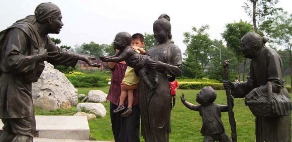 This sculpture is of a child offering an older person some food while a grandmother and another young child watch.  Included in this photo are mother and small child interacting with the sculpture. 